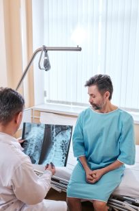 Sad mature patient with cancer sitting on bed and listening to doctors recommendations while doctor viewing x-ray image