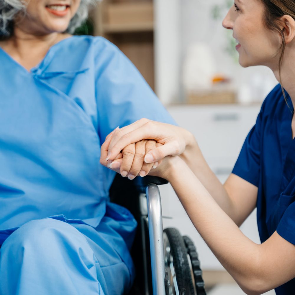 Young nurse helping elderly woman walk in the room, holding his hand, supporting. Treatment and rehabilitation after injury in assisted living facility, senior care
